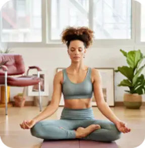 A woman in a serene yoga pose, sitting cross-legged on a mat in a well-lit room with plants and large windows.